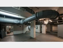 Industrie- oder Technikraum mit großen, sichtbaren Lüftungsrohren an der Decke sowie Betonwänden und -säulen. Im Hintergrund befinden sich eine offene Tür, ein Schaltkasten und weitere technische Anlagen.