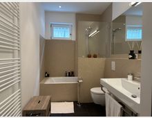 Modernes Badezimmer mit hellen Fliesen an Wänden und Badewanne, kombiniert mit dunklem Boden. Zu den sichtbaren Elementen gehören ein Waschbecken mit Spiegel, eine Toilette, ein Handtuchhalter, ein Heizkörper, sowie ein kleines Fenster mit Tageslicht.