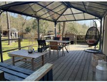 Überdachte Holzterrasse mit Sitzmöbeln, einem hängenden Korbsessel und Blick auf einen Garten mit Bäumen und einer kleinen, dekorativen Metallkuppel im Hintergrund. Im Vordergrund stehen ein Holztisch und eine Milchkanne als Dekoration.