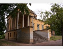 Gelbliches historisches Gebäude mit klassizistischer Architektur, gekennzeichnet durch hohe Säulen an der Vorderseite, eine Freitreppe aus Stein und symmetrisch angeordnete Fenster. Umgeben von Bäumen und Vegetation, steht es in einer parkähnlichen Umgebung.