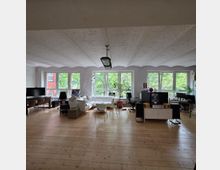 Ein geräumiger, heller Raum mit Holzboden, möbliert mit Schreibtischen, Stühlen, einem Sofa und Zimmerpflanzen. Große Fenster an der Stirnwand bieten Blick auf grüne Bäume und lassen viel Tageslicht hinein.