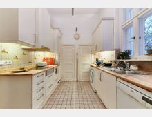 Helle Küche mit weißen Schränken, Holzarbeitsplatten und gefliesten Böden. An den Wänden befinden sich helle Fliesen mit dekorativen Motiven, rechts ist ein großes Fenster, unter dem eine Spüle und einige Pflanzen stehen.