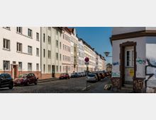 Straßenszene mit mehrstöckigen Wohngebäuden in hellen Farben und roten Ziegelsockeln, die links entlang einer gepflasterten Straße stehen. Vor den Gebäuden parken zahlreiche Autos, während im Vordergrund ein Eckeingang mit Graffiti zu sehen ist.