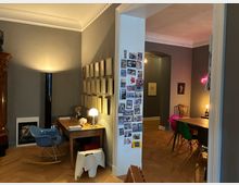 Blick in eine Wohnung mit hohen Decken, Stuckleisten und Parkettboden: links ein kleiner Arbeitsplatz mit Holztisch, Stuhl und Wandrahmen, in der Mitte ein Türrahmen mit Fotocollage. Rechts schließt ein Essbereich mit Holztisch, bunten Stühlen und gedämpfter Beleuchtung an, dahinter ein Flur mit Rundbogen.