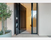 Eingangsbereich eines modernen Wohnhauses mit geöffneter, dunkler Haustür und seitlichen Glasflächen in schwarzem Rahmen. Vor der Tür stehen große Steinfliesen und ein Pflanzkübel mit kleinem Baum; innen ist ein heller Flur mit Deckenspots zu sehen.