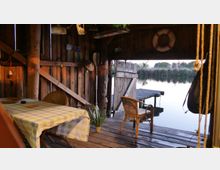 Holzhütte am Wasser mit rustikalem Innenraum, bestehend aus einer Holzterrasse, einem Tisch mit kariertem Tischtuch, geflochtenen Stühlen und dekorativen Elementen wie einem Rettungsring an der Wand. Im Hintergrund führt ein Steg aufs Wasser hinaus, umrahmt von Schilf und Bäumen.