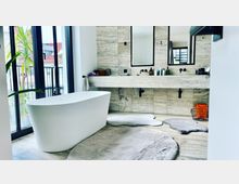 Modernes Badezimmer mit freistehender weißer Badewanne, großflächigen Steinfliesen an Wänden und Boden sowie breitem Waschtisch mit zwei Spiegeln. Große Fenster lassen viel Tageslicht hinein, während Teppiche und dekorative Details wie Pflanzen und Pflegeprodukte den Raum gestalten.