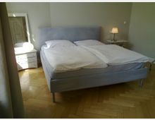 Ein schlicht eingerichtetes Schlafzimmer mit einem Doppelbett mit grauem Polsterkopfteil und weißen Bettdecken, einem Nachttisch mit Lampe rechts und einer Kommode mit Spiegel links. Der Boden ist mit hellem Fischgrätparkett bedeckt, und die Wände sind neutral gestrichen.