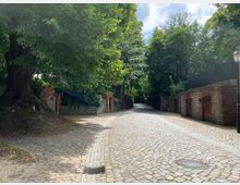 Kopfsteinpflasterstraße, umgeben von roten Backsteinmauern und bewachsenen Toren, führt durch eine bewaldete Umgebung mit großen, schattenspendenden Bäumen. Im Vordergrund links steht ein alter Baumstamm, rechts sind Laternen und verzierte Holztüren in Backsteinfassaden zu sehen.