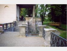 Steinerne Terrasse eines historischen Gebäudes mit Balustraden, kunstvollen schmiedeeisernen Geländern und Treppenaufgang, umgeben von einer grünen Parklandschaft mit Bäumen und einem Holzsteg über einen Bach.
