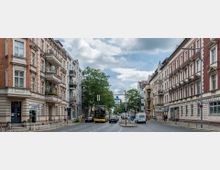 Straßenszene in einer Stadt mit historischen Wohngebäuden auf beiden Seiten, meist aus Backstein und Putzfassaden mit Balkonen. In der Mitte verläuft eine mehrspurige Straße mit Autos, einem gelben Doppeldeckerbus und Begrünung; im Hintergrund sind Bäume und moderne Gebäude zu sehen.