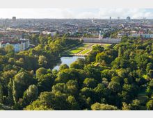 Luftaufnahme eines weitläufigen Schlossparks mit symmetrischen Gartenanlagen, einem großen Schlossgebäude im Barockstil und einem kleinen See, umgeben von dichtem Wald. Im Hintergrund erstreckt sich eine städtische Skyline mit Wohnhäusern und Hochhäusern.