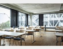 Heller Seminarraum mit modernen Holztischen und Stühlen, großen Fenstern und Blick auf ein benachbartes Gebäude mit Metallstrukturen und Außentreppen. Der Boden ist aus hellem Holz, die Decke besteht aus Akustikplatten.