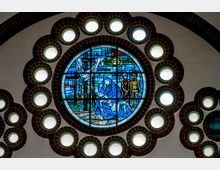 Rundes Kirchenfenster aus Buntglas mit biblischer Darstellung, umgeben von kleineren, kreisförmigen Fenstern mit Ziegelfassungen. Im Mittelpunkt zeigt das Fenster eine Szene mit einer Frau in blauer Kleidung, die einen liegenden Mann hält, vor farbigem Hintergrund.