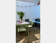 Kleine Terrasse mit Holzboden, grünen Metallstühlen und einem grauen Tisch, daneben ein Ecksofa mit blau und gelb gestreiften Kissen. An der weißen Wand hängt eine Lichterkette, und eine grüne Pflanze steht in einem gestreiften Topf auf dem Tisch.