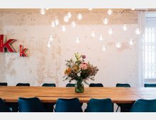 Ein Besprechungsraum mit einem langen Holztisch, umgeben von dunkelblauen Stühlen. Im Mittelpunkt steht eine Vase mit bunten Blumen, und von der Decke hängen runde, leuchtende Lampen; im Hintergrund eine ungleichmäßig verputzte Wand mit großen roten Buchstaben „K“ und „k“.