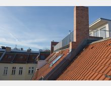 Dächer von Wohngebäuden mit roten Ziegeln und Oberlichtern, ein Ziegelkamin und ein modern gestalteter Balkon im oberen Bereich des Bildes, im Hintergrund blauer Himmel.