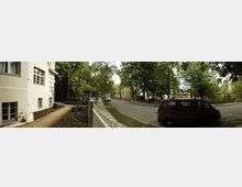 Straßenszene in einem ruhigen Wohngebiet mit gepflastertem Weg, flankiert von einer weißen Hausfassade links, Bäumen und Grünflächen. Ein roter Van parkt rechts am Straßenrand; im Hintergrund sind weitere Gebäude und Bäume zu sehen.