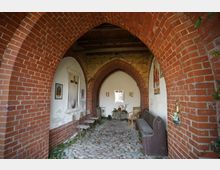 Überdachter Gang oder kleine Kapelle mit spitzbogigen Backsteinarkaden und Kopfsteinpflasterboden, an den weiß verputzten Wänden hängen religiöse Bilder. Entlang der Seiten stehen einfache Stühle und eine Holzbank; am Ende befindet sich ein kleiner Altar mit Blumen und Kerzen.