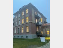 Ein mehrstöckiges Wohngebäude mit rosa Putzfassade und weißen Fensterrahmen, die von innen beleuchtet sind. Vor dem Gebäude befindet sich eine Wiese und ein Weg mit einem Schild, das die Adresse "Mariendorfer Weg 30" angibt.