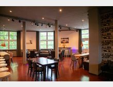 Moderner Innenraum eines Cafés oder Veranstaltungsraums mit roten Böden, großen Fenstern, grauen Vorhängen und verschiedenen Sitzbereichen mit Holztischen und gepolsterten Stühlen. Rechts befindet sich eine Wand aus Naturstein, und von der Decke hängen dezente Beleuchtungskörper.