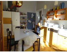 Helle Küche mit moderner Einrichtung, darunter ein weißer Esstisch mit Stühlen, eine goldene Hängeleuchte und offene Holzregale an der Wand. Die Küchenschränke und Arbeitsflächen sind aus Holz und Weiß, während eine große Kühl-Gefrierkombination und Haushaltsgeräte sichtbar sind.