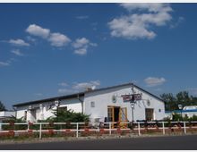 Ein einstöckiges Gebäude mit weißer Fassade und braunen Fensterrahmen, das als Restaurant dient. Vor dem Gebäude befinden sich eine Terrasse mit Tischen und Stühlen, Blumenkästen sowie eine niedrige Absperrung mit rot-weißen Elementen; im Hintergrund ist ein klarer blauer Himmel sichtbar.