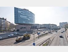 Moderne Bürogebäude mit einer geschwungenen Glasfassade, das auf einem massiven Unterbau ruht, an einer mehrspurigen, stark befahrenen Straße mit danebenliegenden Wohngebäuden und einer erhöhten Verkehrsführung. Im Vordergrund ist ein kleiner Lastwagen auf der Straße sichtbar.