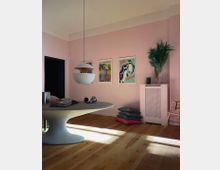Ein heller Wohnraum mit hellrosa Wänden, einem runden Tisch mit organischem Design in der Mitte und einer hängenden, modernen Pendelleuchte. An der Wand hängen bunte Poster, daneben befinden sich ein Heizkörper mit Abdeckung, eine Vase mit grünen Pampasgraszweigen und auf dem Boden liegen Kissen auf dem Holzboden.