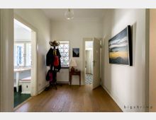 Ein heller Flur mit Holzboden, links ein offener Durchgang zu einer Sitzecke mit Fenstern und Sprossenverglasung. An der rechten Wand hängt ein großes Gemälde, in der Mitte stehen eine Garderobe mit Jacken und Taschen sowie ein kleiner Tisch mit Lampe. Im Hintergrund führt eine Tür zu einem weiteren Raum mit schwarz-weißen Fliesen.