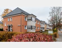 Mehrfamilienhäuser im Stil der 1920er- oder 1930er-Jahre mit orange- und beigefarbenen Fassaden, großen Fenstern und Ziegeldächern. Im Vordergrund Gehweg, Herbstbäume und eine Hecke mit roten Blättern.