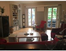 Wohnzimmer mit Holzboden, einem weißen Couchtisch und roten Sitzmöbeln. Im Hintergrund eine große Fensterfront mit Glastüren, die auf eine Terrasse mit einem Grill und Pflanzen führen; links befindet sich ein Regal mit Büchern und Gegenständen.