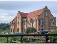 Backsteingebäude im Stil eines traditionellen Landhauses mit Ziergiebeln und Fensterläden in Schwarz-Weiß. Im Vordergrund eine Gartenanlage mit Blumen, Holzzaun und Laternen; dahinter parkende Autos.
