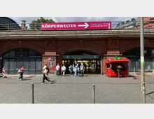 Eingang zum S-Bahnhof Hackescher Markt in Berlin, unter einem historischen Ziegelbogenbau mit darüber liegendem Viadukt. Vor dem Eingang stehen Passanten, und rechts befindet sich ein roter Verkaufsstand in Form einer Erdbeere.