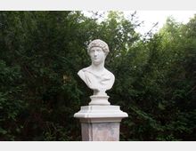 Büste einer klassischen Statue aus weißem Marmor auf einem Sockel aus Stein, umgeben von dichtem grünen Laub und Bäumen im Hintergrund.