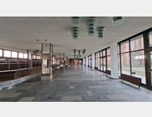 Große, leere Innenhalle mit Steinboden, Rasterdecke und mehreren zylindrischen Deckenleuchten sowie weißen Stützen. Rechts verläuft eine lange Glasfassade mit Blick auf Backsteinbauten und einen Hof; links sind eine Theke bzw. Garderobenanlage und Backsteinwände zu sehen.