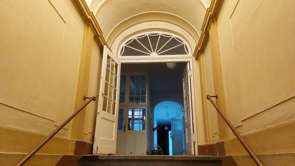 Treppenaufgang in einem historischen Gebäude mit weiß gestrichenen Wänden, verzierten Bögen und offenen Doppeltüren. Dahinter sind weitere Glastüren und ein Flur mit blauer Beleuchtung sichtbar.