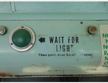 Nahaufnahme einer alten, hellgrünen Metalloberfläche mit einem grünen Leuchtknopf links, der von einem Metallring umgeben ist. Daneben befindet sich der Text „WAIT FOR LIGHT“, gefolgt von einer Anleitung zum Öffnen einer Tür.