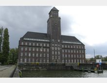 Ein großes Backsteingebäude mit einem zentralen Turm, an dem eine Uhr angebracht ist, steht am Ufer eines Kanals. Das Gebäude hat viele symmetrische Fenster und ein dunkles Schieferdach, umgeben von Bäumen und einer gepflasterten Straße.