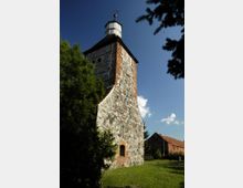 Turm einer historischen Kirche aus Feldsteinen mit Fachwerkelementen und einer hölzernen Laterne an der Spitze. Im Vordergrund Bäume und Rasen, im Hintergrund ein weiteres Gebäude mit Ziegeldach.