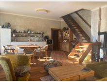 Rustikales Wohnzimmer mit offener Küche, Holzboden und einer hölzernen Treppe, die nach oben führt. Der Raum enthält eine Sitzecke, einen runden Esstisch, offene Regale mit Geschirr sowie eine Kombination aus modernen und rustikalen Möbeln.