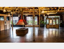 Großzügiger Raum mit industriellem Design, gekennzeichnet durch freiliegende Metallträger, Lüftungsrohre und Hängelampen. Im Zentrum befindet sich ein großer, moderner Kamin mit gemauerter Basis und roter, pyramidenartiger Haube; im Hintergrund große Fenster und Türen, dahinter Sicht auf Pflanzen und Außenbereich.