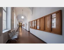 Langer, heller Flur eines historischen Gebäudes mit weiß gestrichenen Wänden und einem gewölbten Deckenbogen. Auf der linken Seite befinden sich große, rechteckige Fenster und einige Stühle, auf der rechten Seite hölzerne Schaukästen mit Glasscheiben.