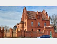 Backsteinbau im neugotischen Stil mit zahlreichen spitzbogigen Fenstern und Zinnen entlang des Dachs, umgeben von einer gemauerten Ziegelmauer mit Torbogen. Vor dem Gebäude steht ein Auto am Straßenrand, und die Bäume im Hintergrund sind kahl.