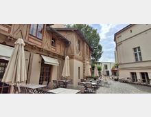 Gepflasterter Innenhof mit Café-Bestuhlung und mehreren geschlossenen Sonnenschirmen, flankiert von einem Fachwerkgebäude mit großen Fenstern und einem hellen Putzbau. Im Hintergrund stehen Bäume und weitere niedrige Gebäude, wodurch der Hof wie ein kleiner, geschützter Platz wirkt.