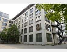 Mehrstöckiges weißes Gebäude mit großen rechteckigen Fenstern und einer schwarzen externen Metalltreppe auf der rechten Seite. Im Vordergrund befinden sich ein gepflasterter Hof und Bäume, die teilweise das Gebäude verdecken.