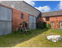 Backsteinbau mit Metalltoren und einem angrenzenden Gebäude mit Ziegeldach. In einem begrünten Hof steht eine Schaukel mit Sonnendach, und im Vordergrund liegt eine Plane auf dem Boden.