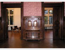 Holzvertäfelter Innenraum mit zwei offenen Türen, die in benachbarte Räume mit großen Fenstern führen. Die Wände sind mit rötlich gemusterten Tapeten gestaltet, und der Boden besteht aus dunklem Parkett. Im Vordergrund befindet sich ein dekorativer hölzerner Wandaufsatz.