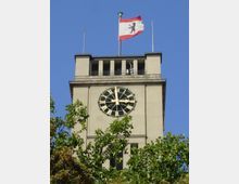 Turm eines Gebäudes mit einer großen, runden Uhr mit römischen Ziffern auf der Vorderseite, darüber weht die Flagge von Berlin. Im Vordergrund sind grüne Baumkronen zu sehen, der Himmel ist blau und wolkenlos.