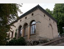 Historisches Gebäude mit rechteckigem Grundriss, hellbrauner Fassade und drei großen Rundbogenfenstern auf der Vorderseite. Die Architektur weist klassische Elemente wie Gesimse und Pilaster auf, umgeben von Bäumen und Natursteinmauern.